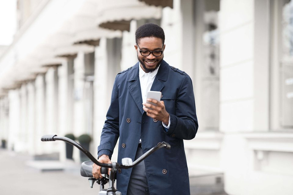 A smiling man in a coat checks his phone while walking his bicycle along a city street